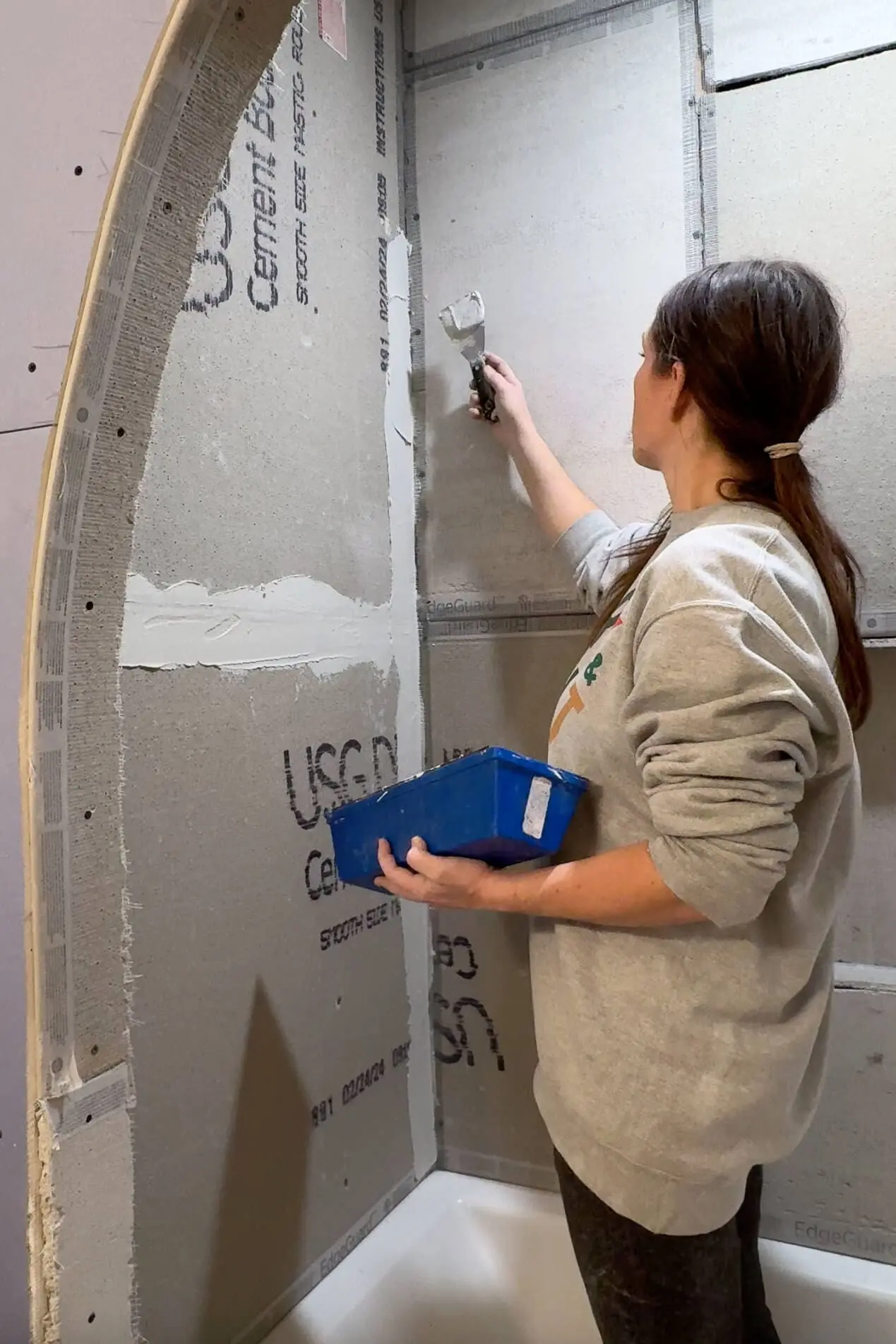 A woman applying thinset mortar with a putty knife along the taped seams of cement boards for a tile tub surround. She holds a blue tray filled with compound in her other hand, smoothing the surface as part of the renovation process.